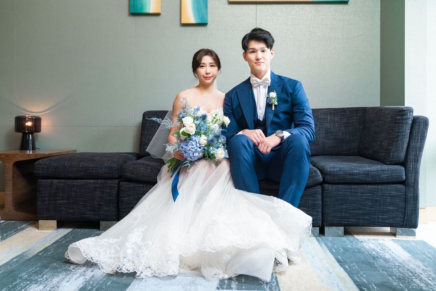 Newlyweds pose together on a modern gray couch, with bride in white gown holding blue bouquet and groom in navy suit.
