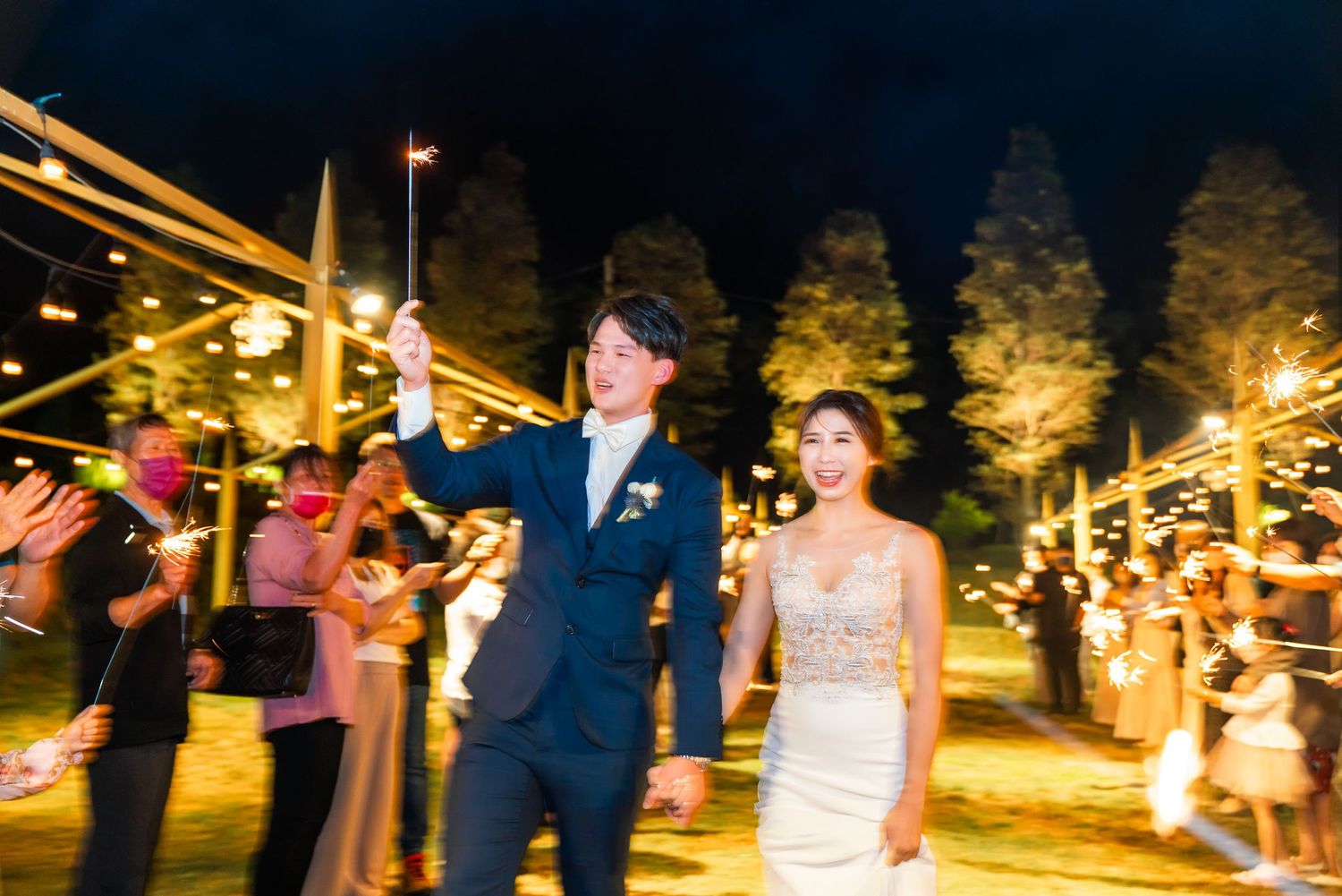 A couple celebrates at an outdoor evening wedding reception under string lights and tall trees.