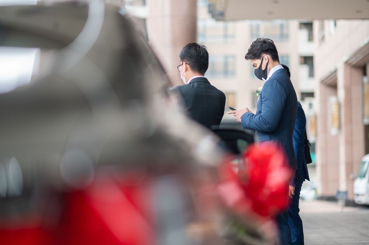 Two businessmen in suits stand on a sidewalk while looking at mobile devices with a blurred red object in foreground.