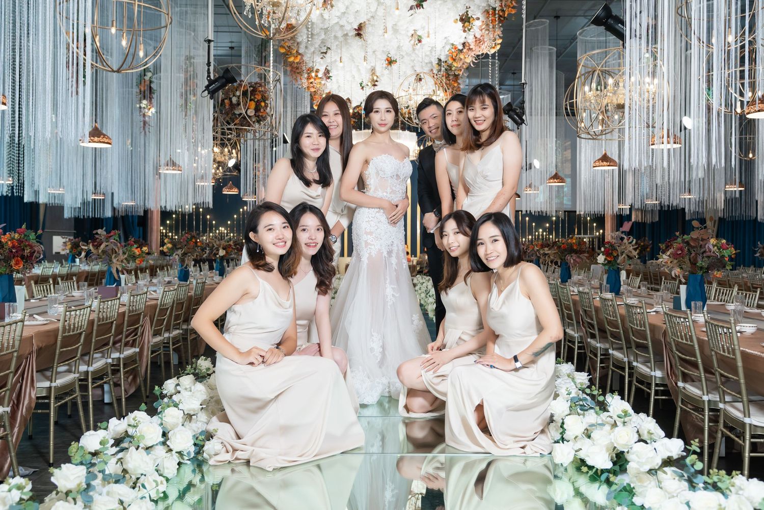 Elegant wedding party in blush pink dresses pose inside luxurious venue decorated with crystal chandeliers and white florals.
