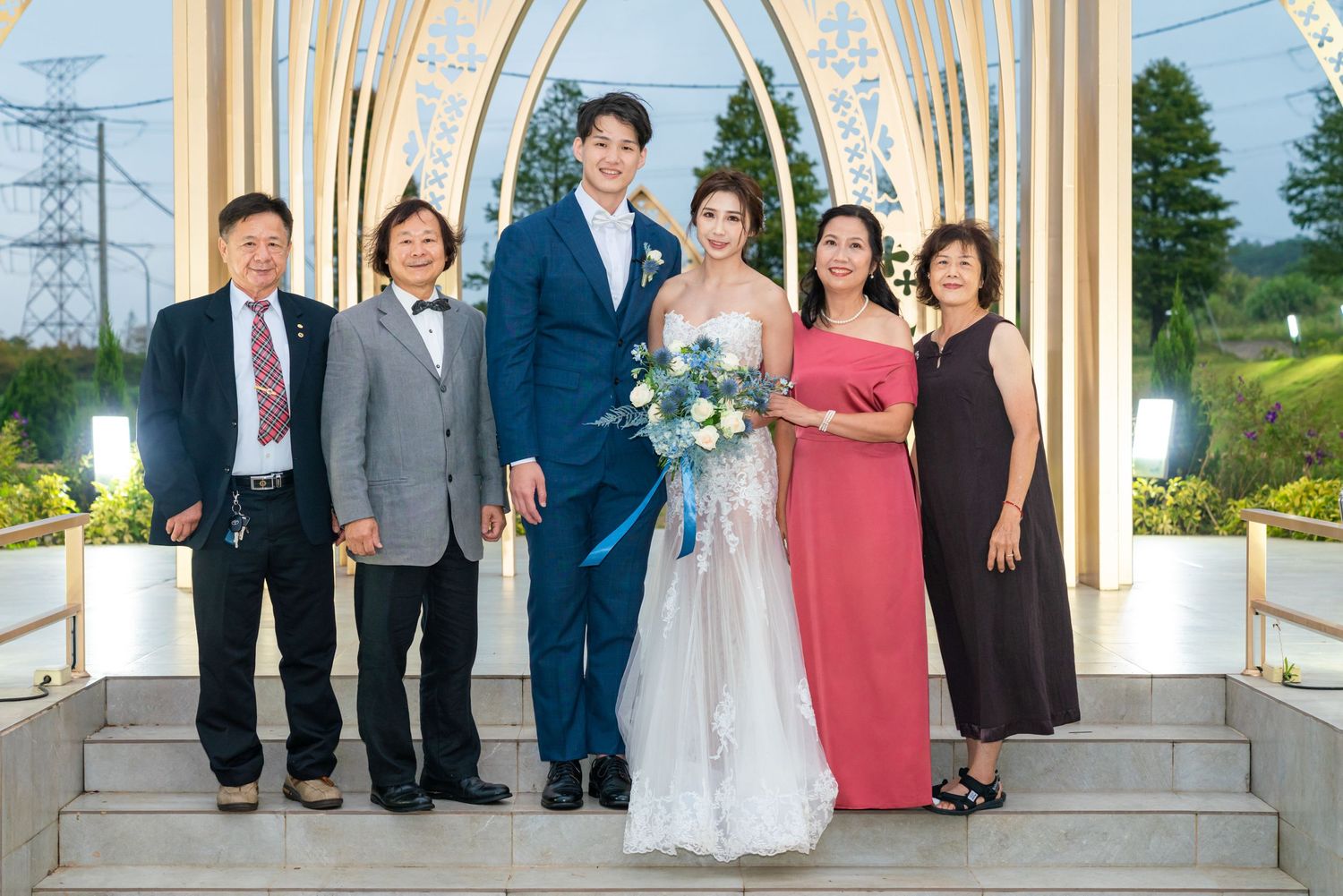 Wedding party poses under an elegant white archway outdoors during a formal celebration ceremony.