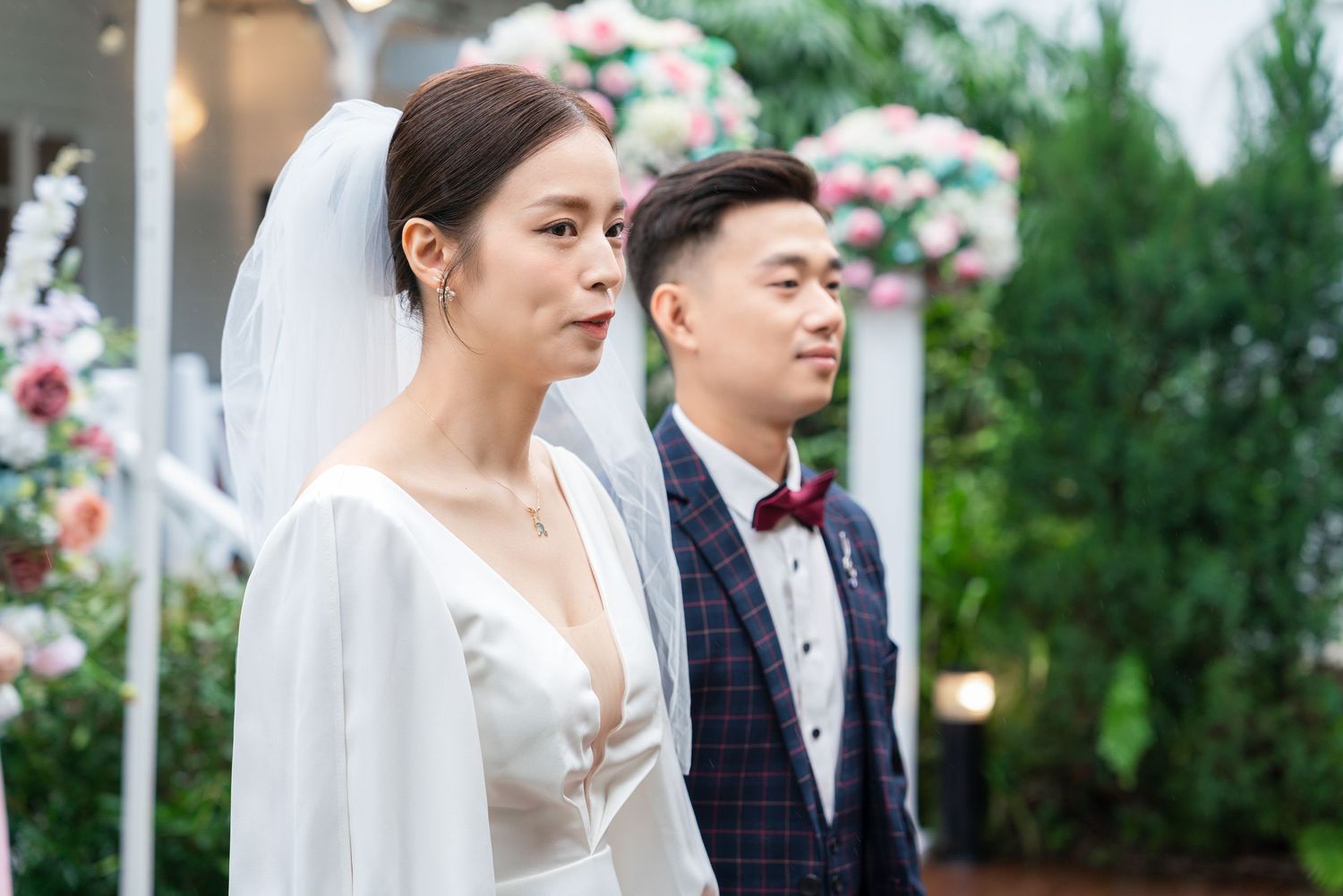 A couple in formal wedding attire stands outdoors with floral decorations at an outdoor ceremony venue.