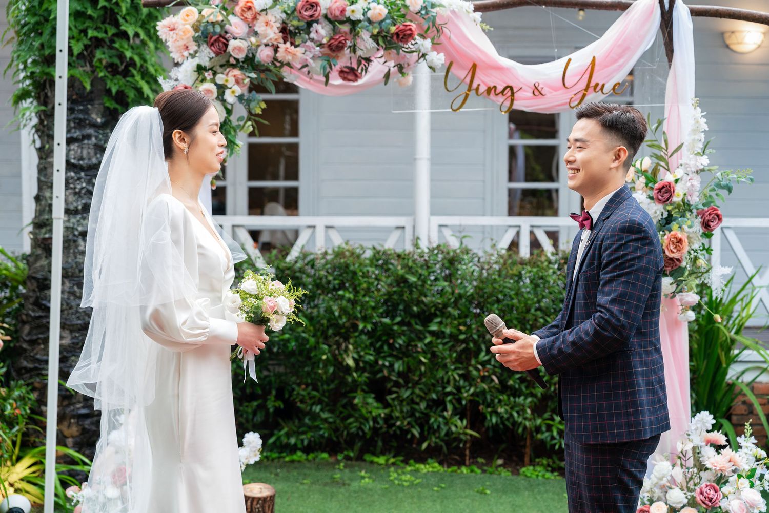 Wedding couple sharing vows under floral arch with pink and white roses during garden ceremony.