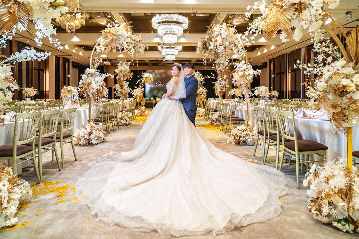 Luxurious ballroom wedding reception with ornate floral arrangements, crystal chandeliers and a sweeping white cathedral train gown.