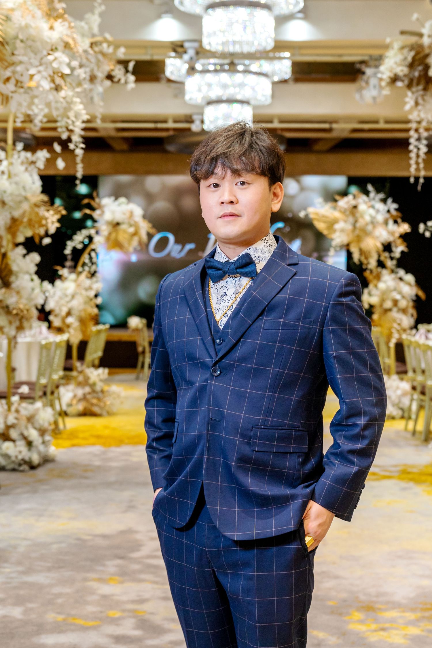 A formal event with a navy checkered suit, bow tie and decorative floral arrangements with chandeliers in an elegant venue.