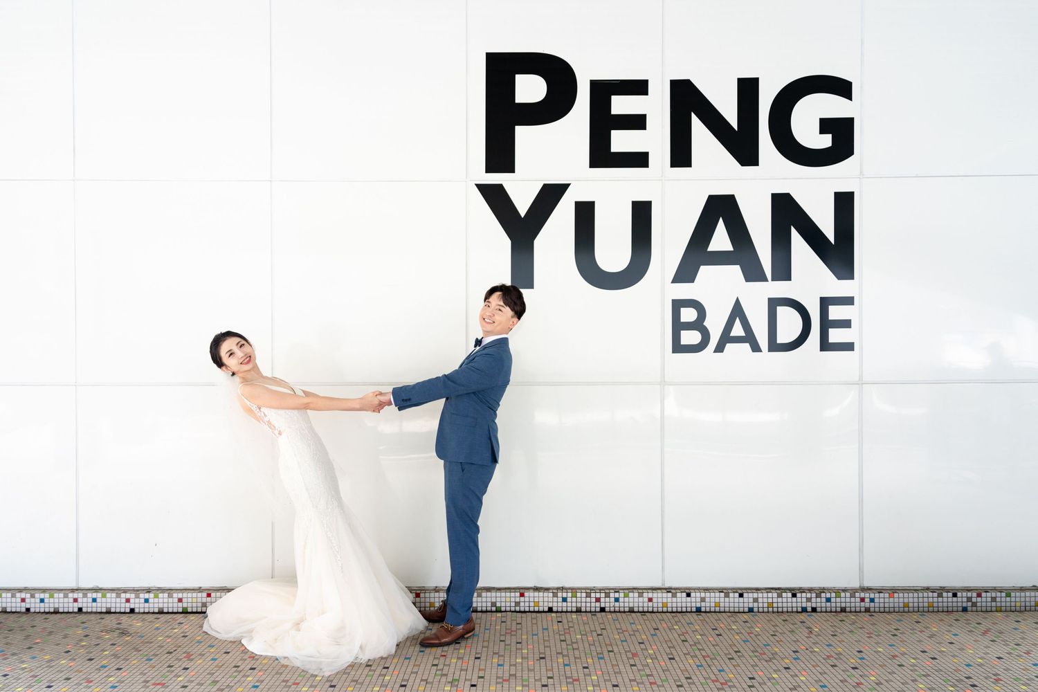 A newlywed couple poses playfully against a white wall with black text reading 'PENG YUAN BADE'.