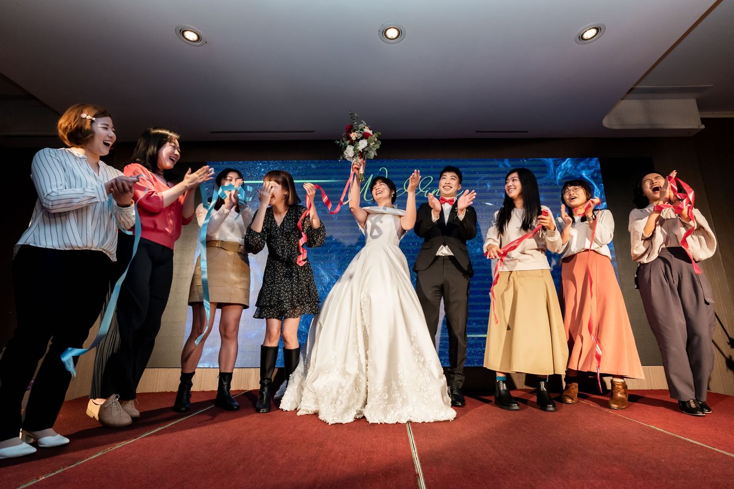 A bride in a white gown holds her bouquet triumphantly surrounded by wedding guests on a red carpet.