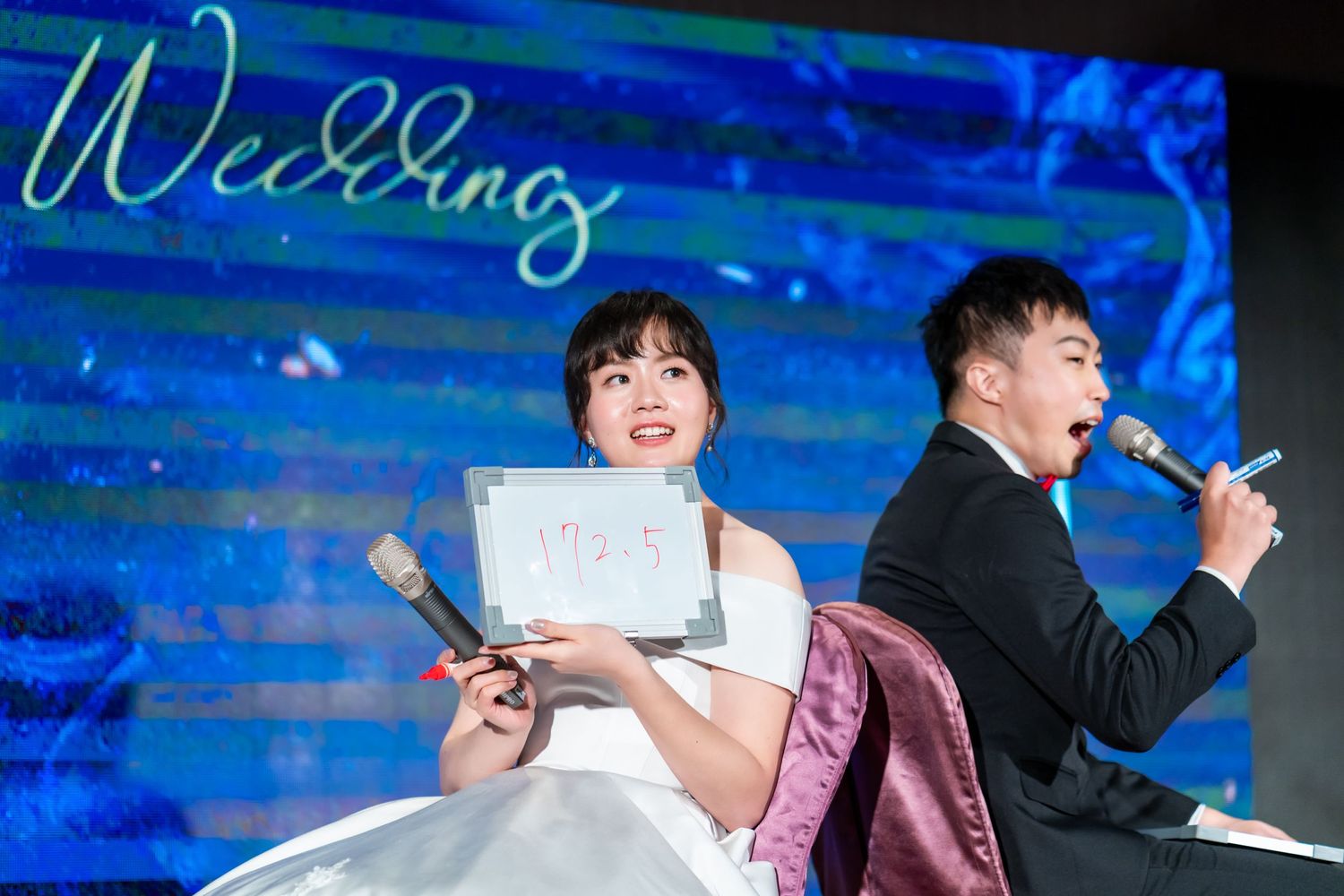 Couple holds white cards while performing at their wedding reception against a blue backdrop.