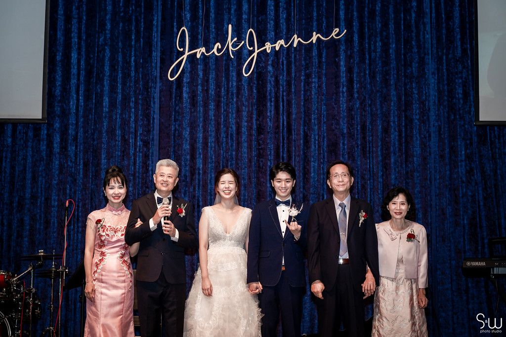 Wedding party group photo against blue curtain backdrop with illuminated signage.