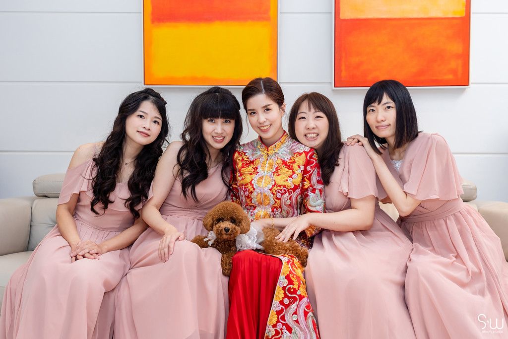 Bridesmaids in pink dresses pose with bride in traditional red qipao dress against colorful wall art.