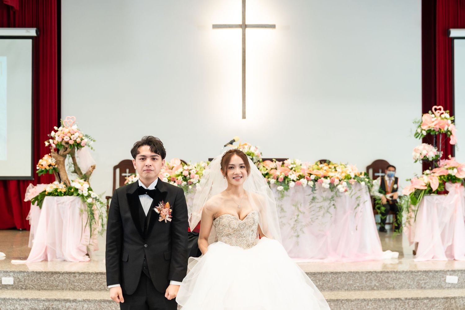 A newlywed couple stands before an illuminated cross in a decorated church sanctuary.