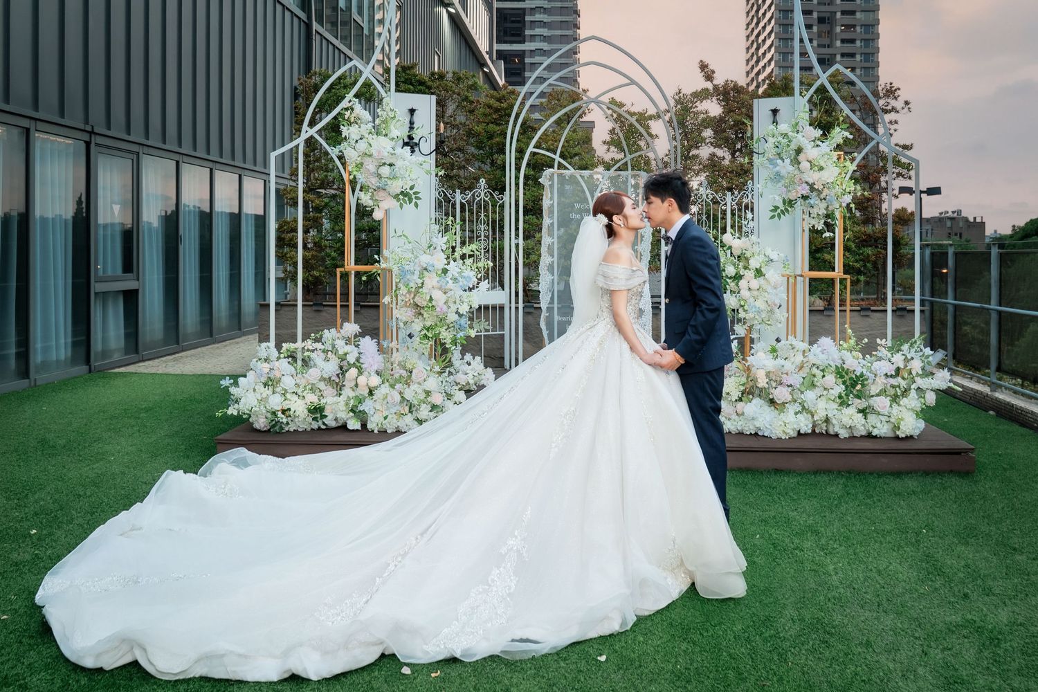 Couple shares romantic kiss in wedding attire beside floral arch on rooftop garden venue.
