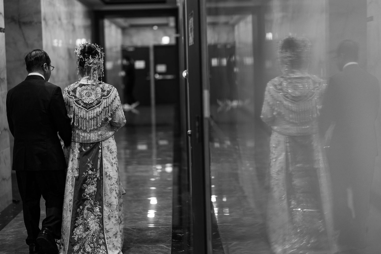Black and white photo of a bride in an ornate traditional Chinese wedding gown walking beside a groom in a suit, reflected in a glass wall.