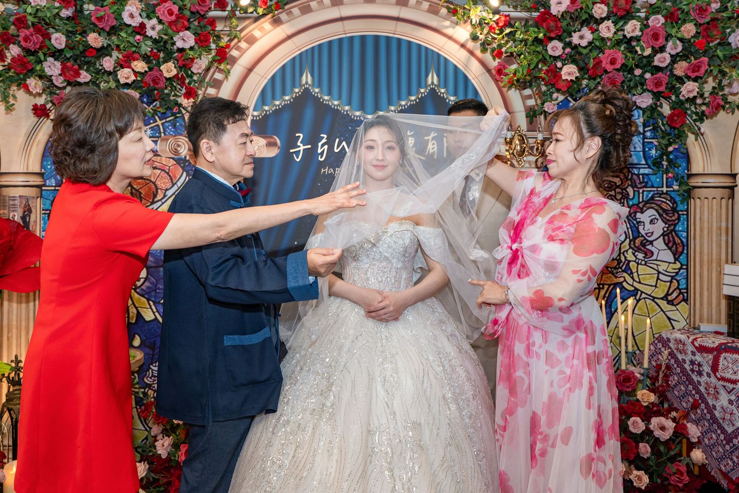 A bride in a white gown has her veil adjusted by family members in a floral wedding ceremony setting.
