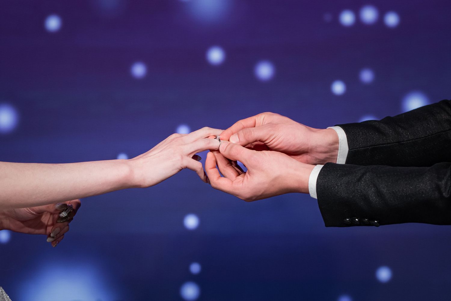 Hands exchanging a ring in a romantic proposal scene with glowing blue bokeh lights in the background.