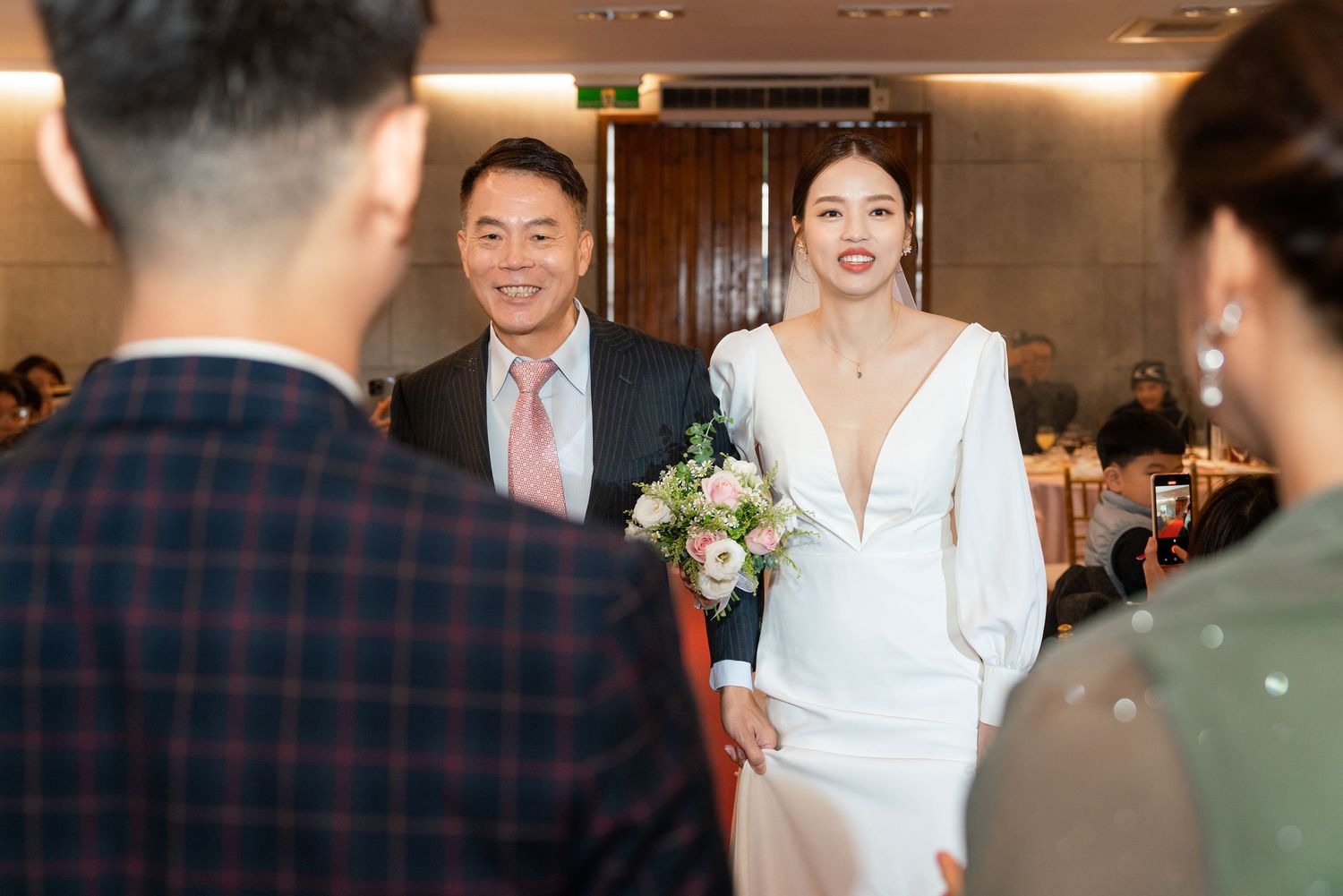 A wedding celebration scene showing a couple at an indoor venue with guests and elegant decor.