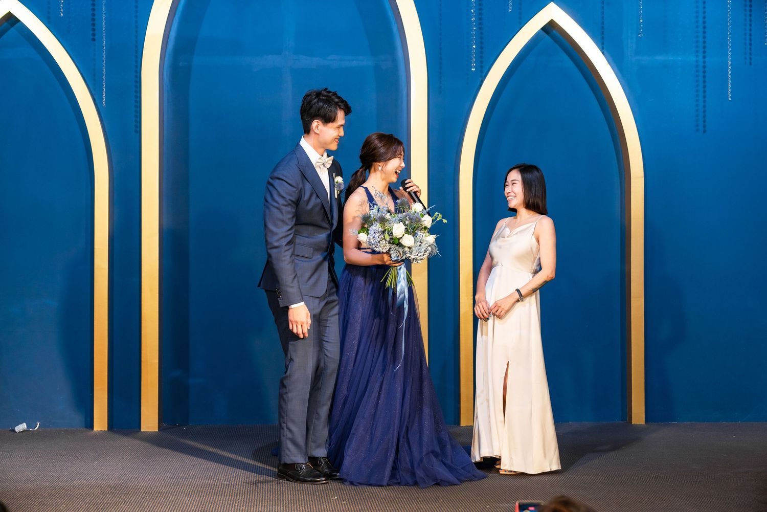 Three people in formal attire standing against a blue wall with gold arched architectural details in an elegant setting.