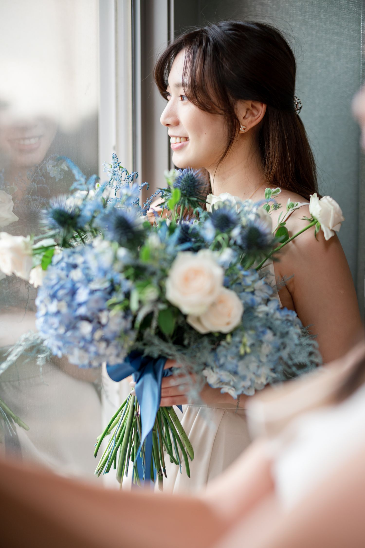 Beautiful bridal bouquet with blue hydrangeas, delphinium, and peach roses tied with a blue ribbon by a window.