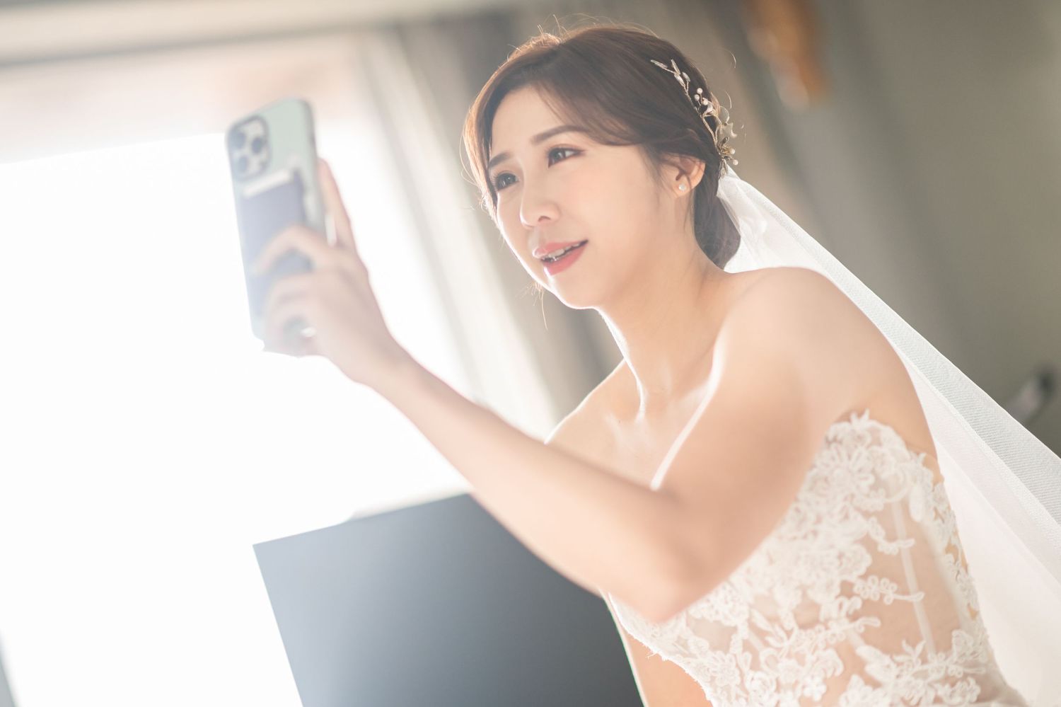 A bride in a white lace wedding dress takes a selfie on her phone while wearing a long flowy veil.