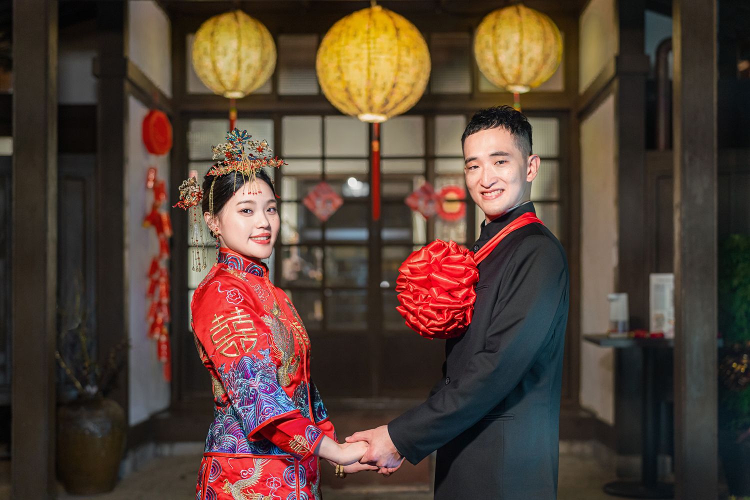 A couple in traditional Chinese wedding attire poses beneath yellow lanterns and red decorations.