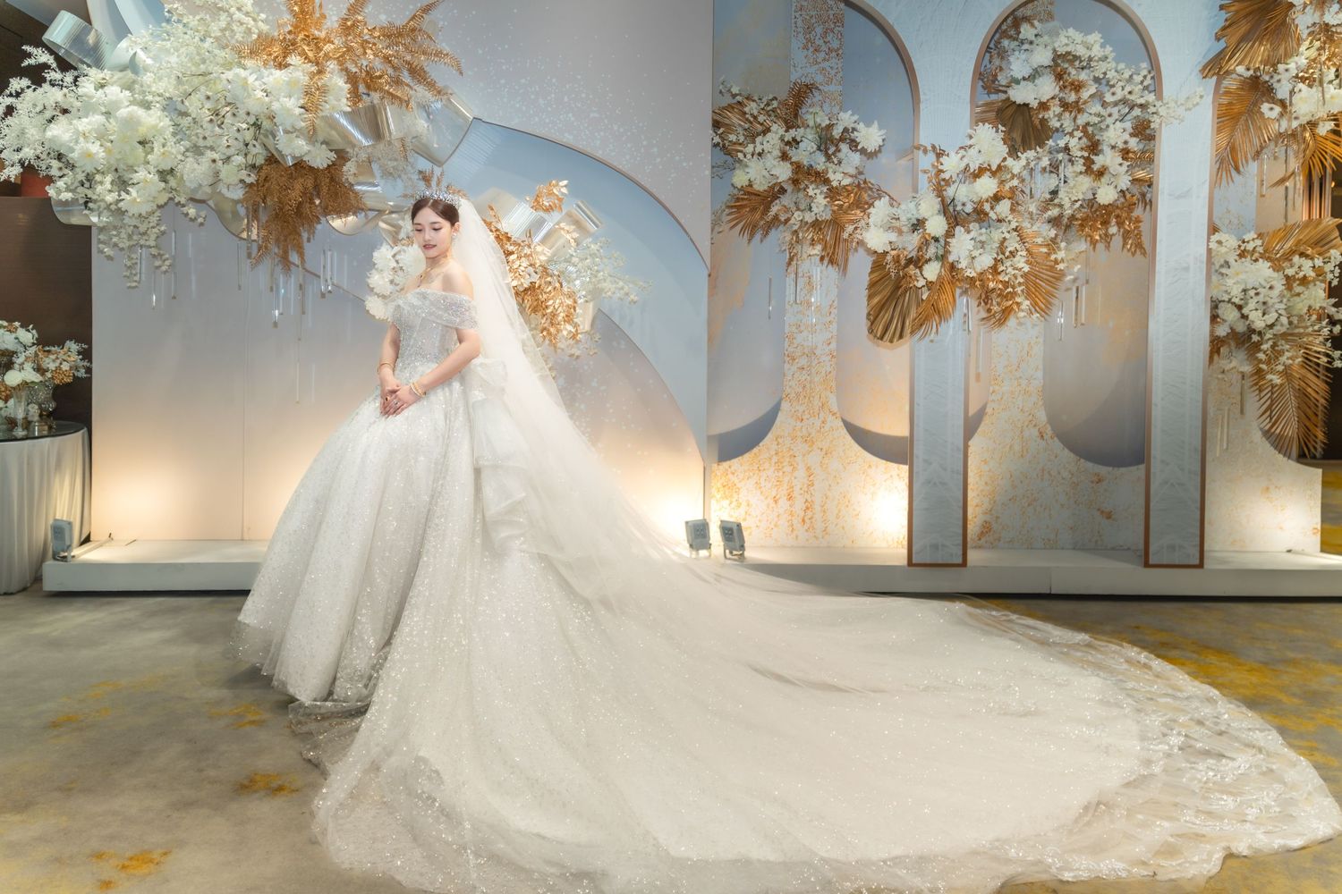 A stunning white wedding gown with elaborate lace details and long cathedral train in an elegant venue with floral decor.