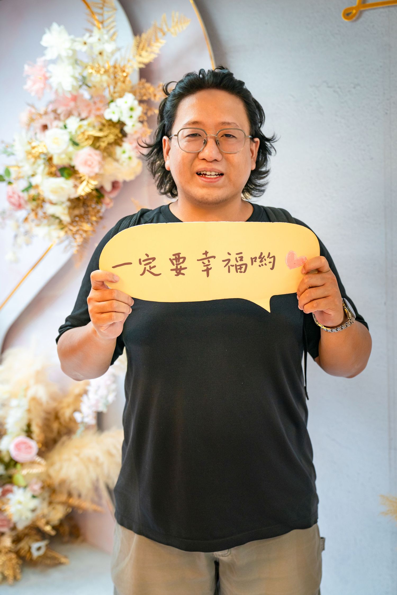 A person in a black t-shirt holds up yellow speech bubble signs with Chinese text against a floral backdrop.