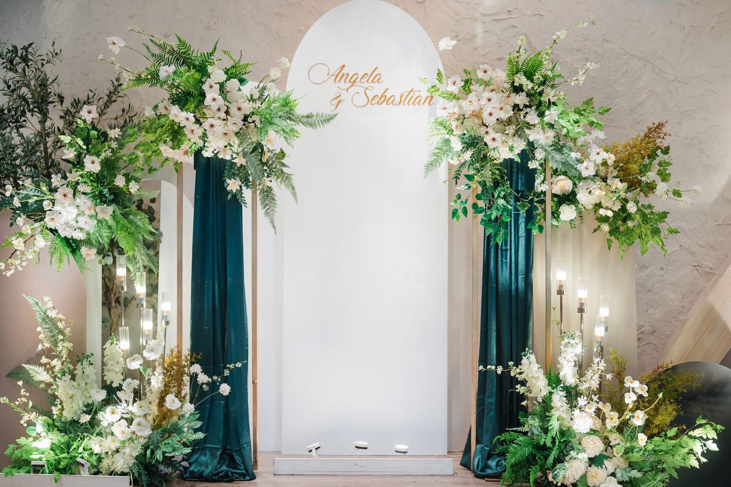 Elegant wedding arch with white flowers, greenery and emerald green drapes against a white stone wall at an indoor venue.