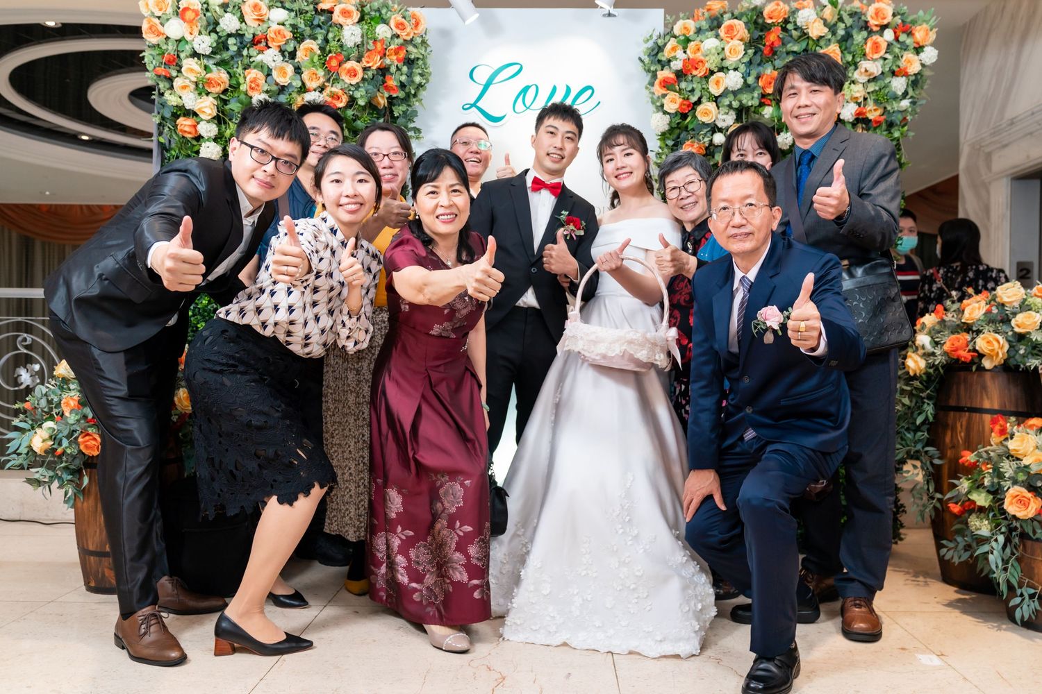 Joyful wedding celebration group photo with guests making fun poses in front of floral backdrop.