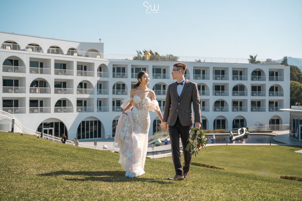 Newlyweds walk together on manicured lawn with white Mediterranean hotel in background.