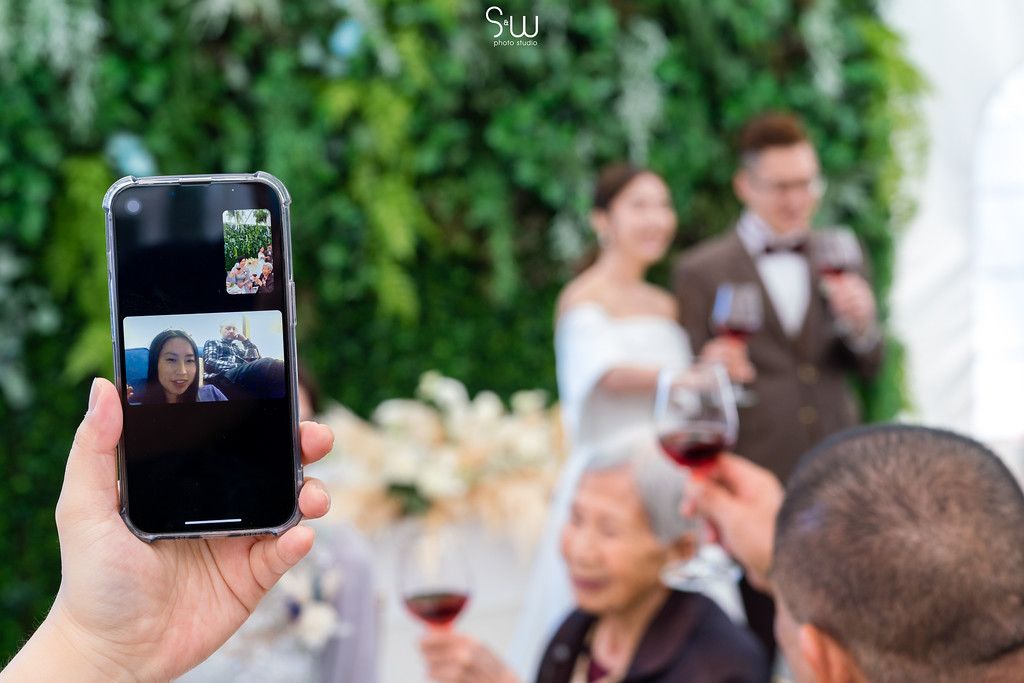 Hand holds phone taking video call during outdoor wedding reception.
