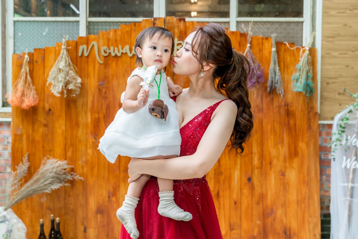 Red gown-clad figure holds a small child in white dress against rustic wooden backdrop.