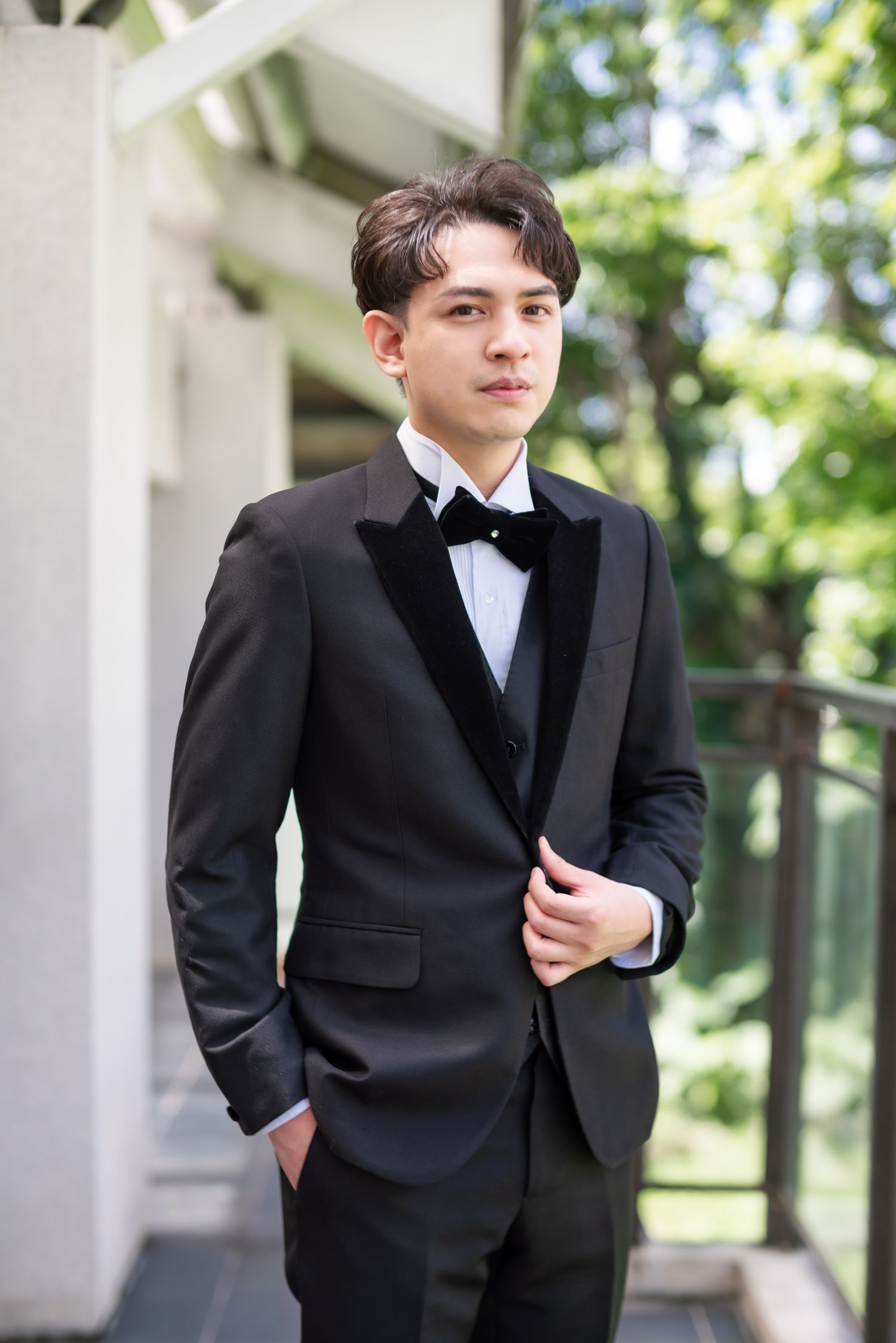 Groom in classic black tuxedo and bowtie poses outdoors against natural greenery background.