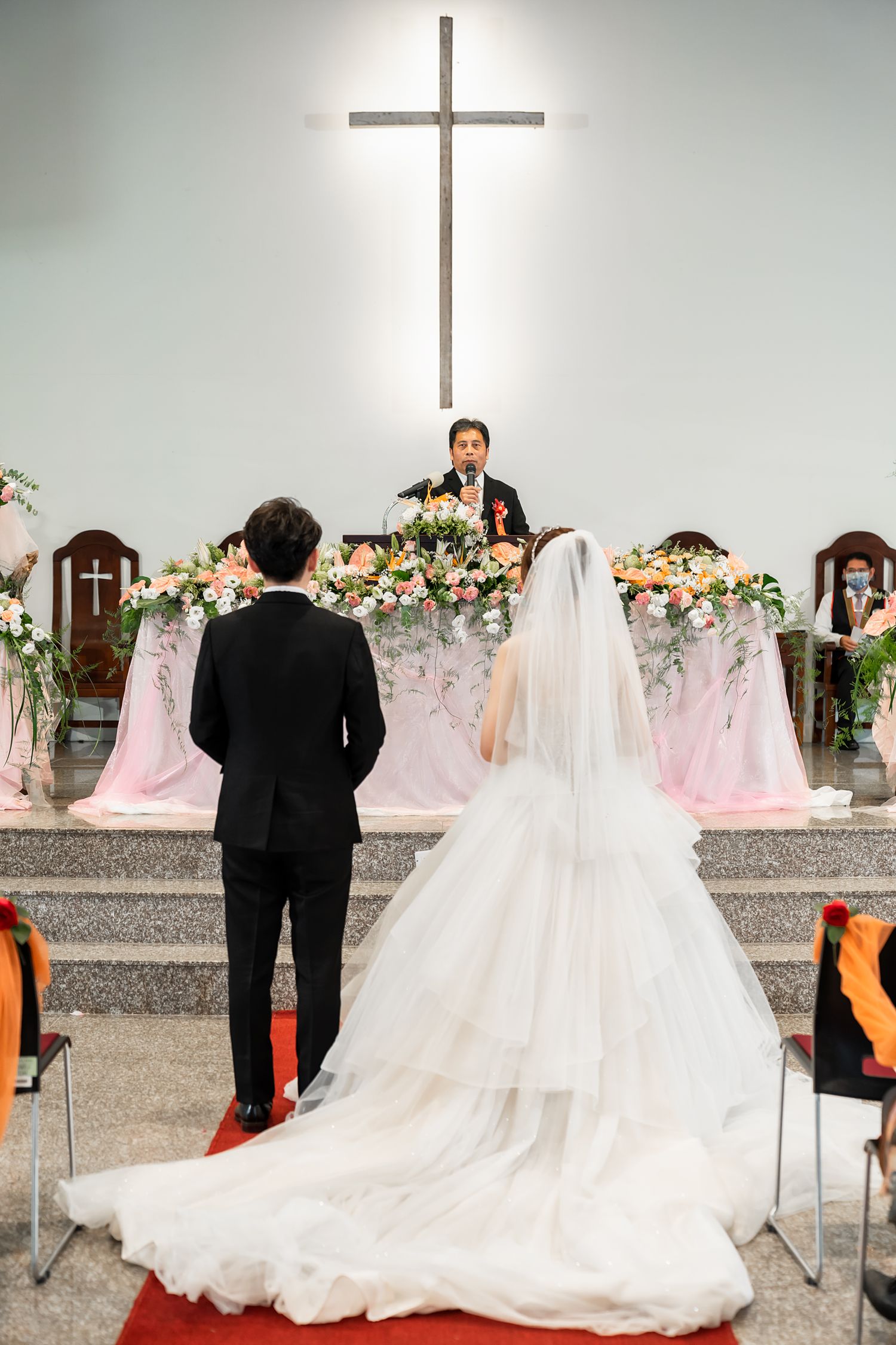 Wedding ceremony with long train dress and illuminated cross above floral-decorated altar.