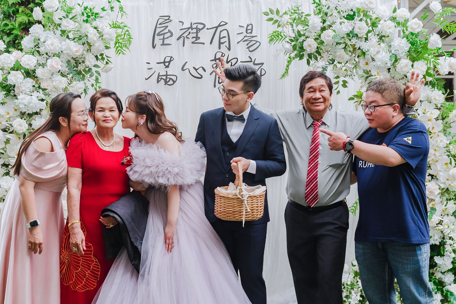 A festive wedding celebration with attendees posing in front of a floral backdrop at an outdoor garden party.