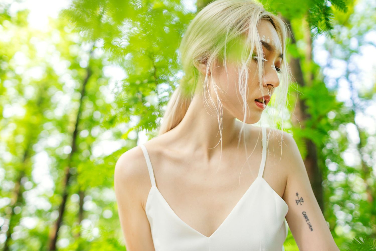 A person in a white camisole poses outdoors in a sun-dappled forest setting among bright green foliage.