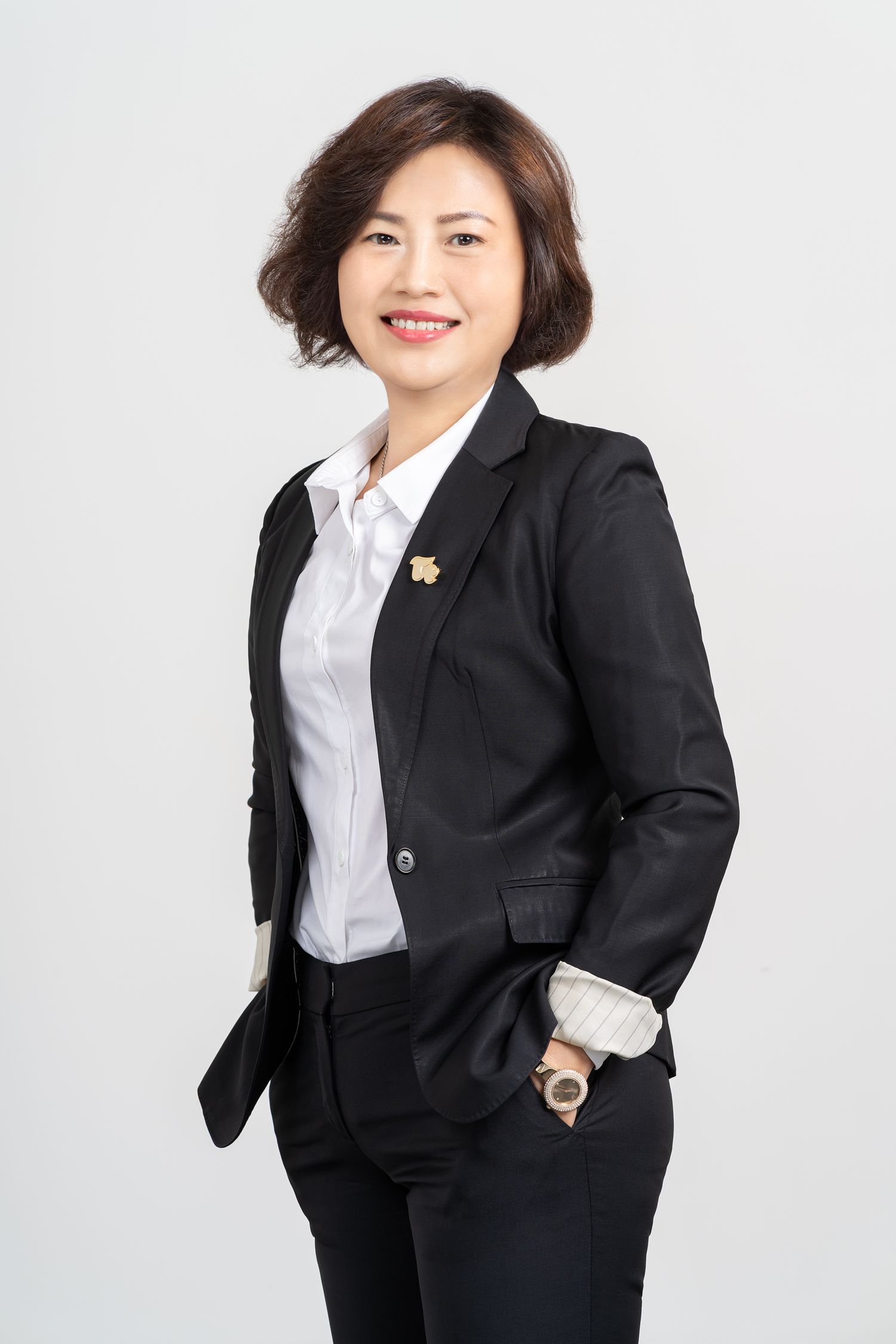 Professional corporate headshot of person in black blazer suit and white button-up shirt against light background.