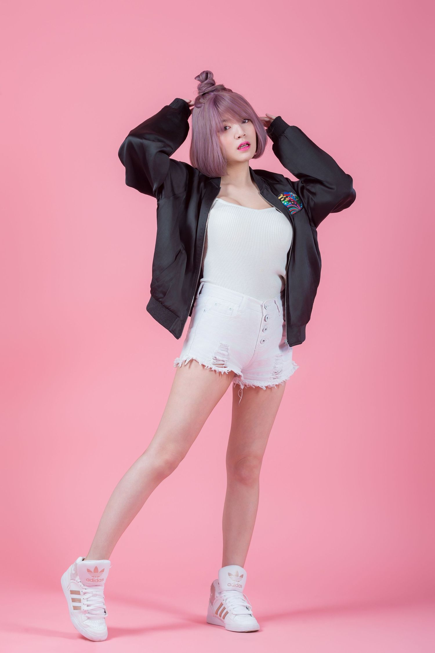 A fashion model poses playfully in an oversized black jacket, white top, denim shorts and white sneakers against a pink backdrop.