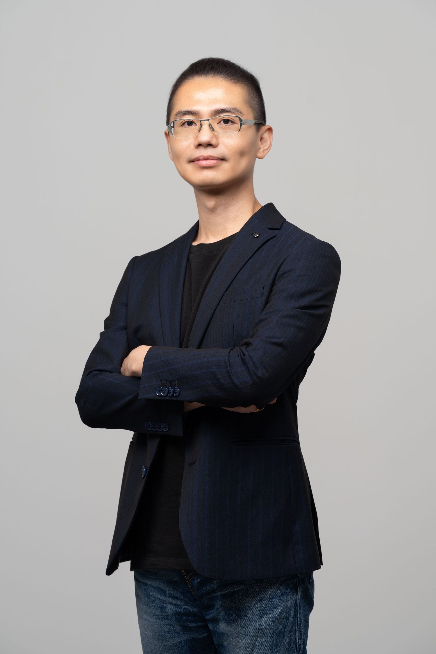 Professional portrait of someone wearing glasses and a navy blazer with arms crossed, against a gray background.