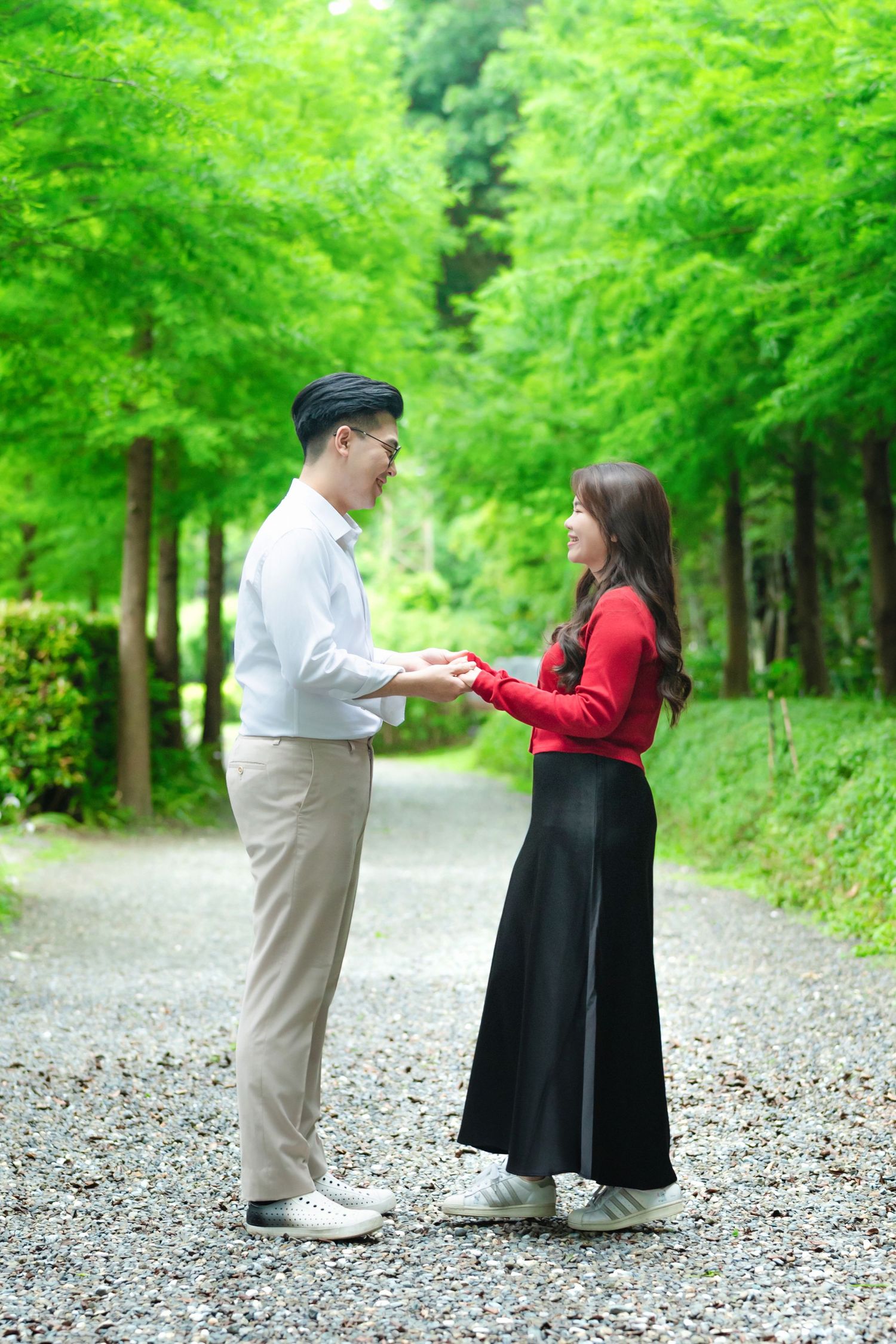 A couple stands together on a tree-lined gravel path surrounded by vibrant green foliage.