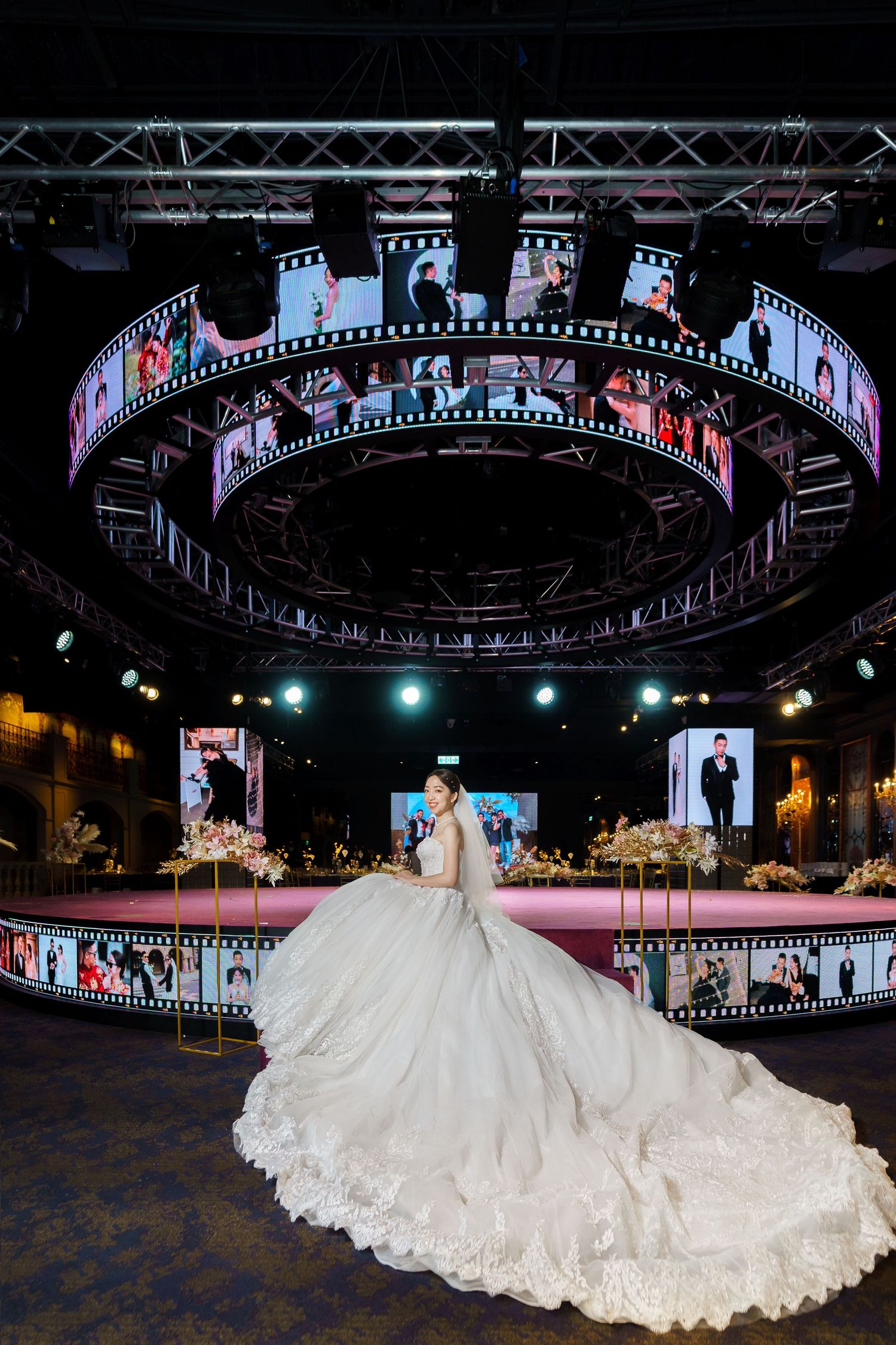 A bride in an elegant white ballgown stands under a circular LED display showing wedding photos at a dark venue.