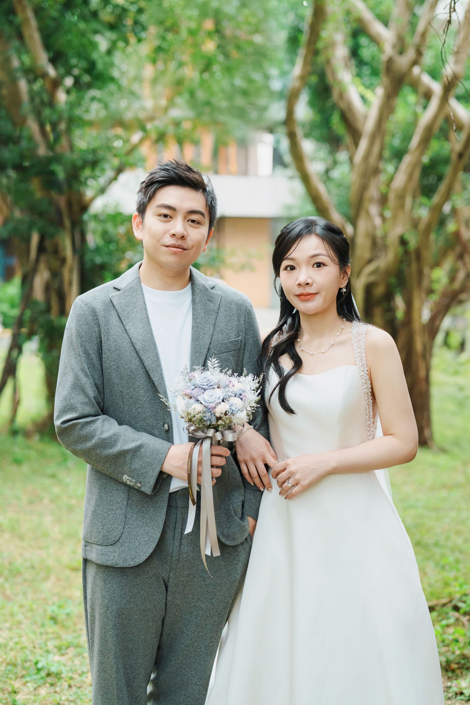 A couple poses together in an outdoor garden setting wearing wedding attire with grey suit and white dress.