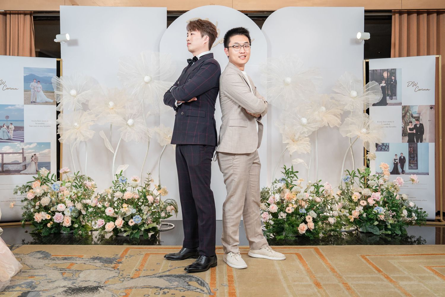 Two men in formal suits stand back-to-back at an elegant wedding venue decorated with flowers.