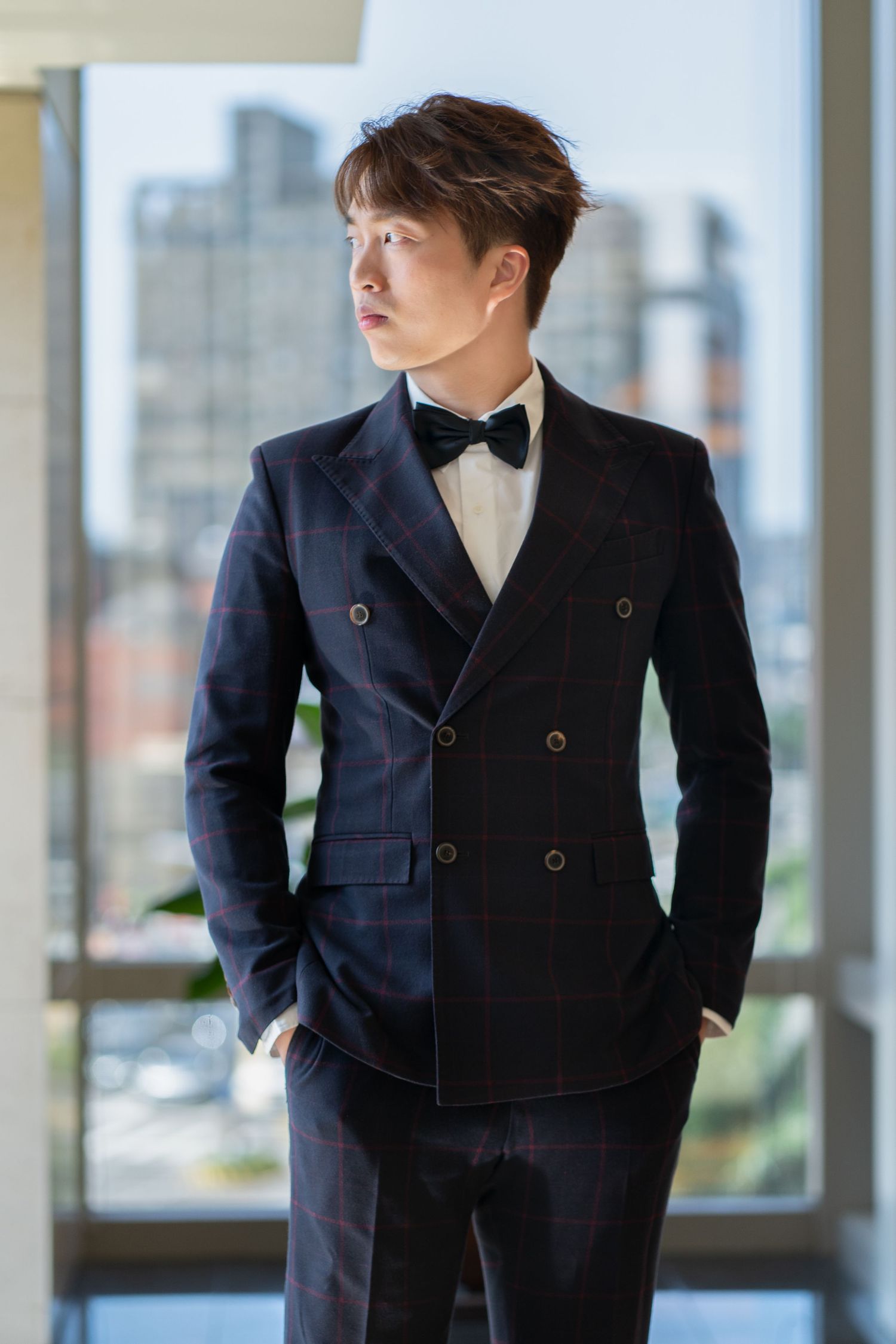 Young person in dark plaid double-breasted suit with bow tie standing by window with city view.