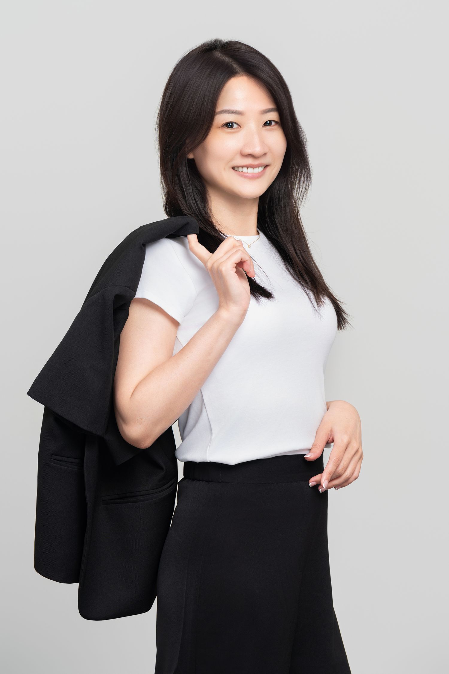 Smiling Asian businesswoman in white top and black skirt holding jacket over shoulder on gray background.