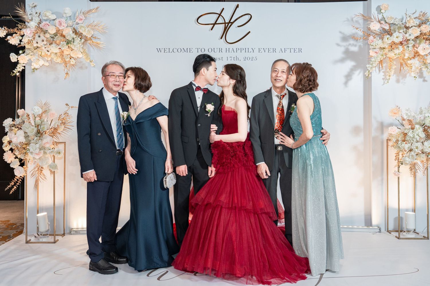 Formal event photo with couples posing in front of decorated backdrop, featuring an elegant red ballgown.