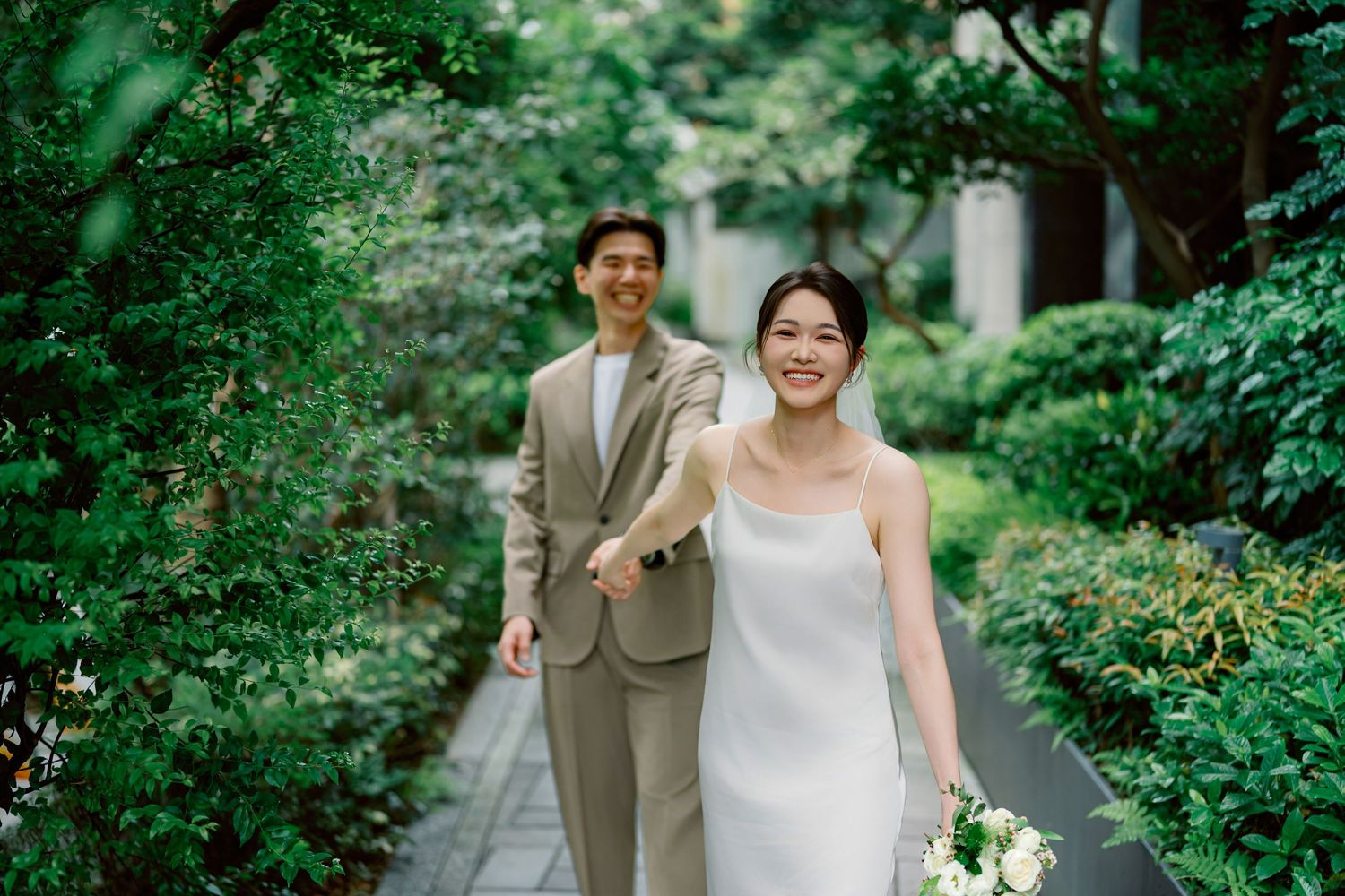 Happy couple walking through lush garden pathway surrounded by greenery on their wedding day.