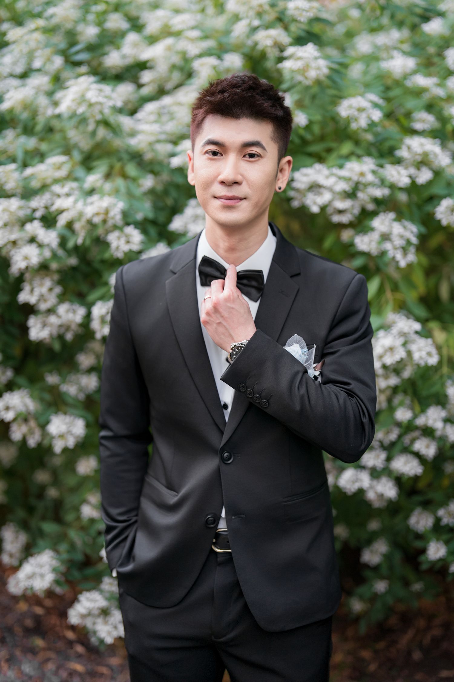 A person in a black suit and bowtie poses playfully against a backdrop of white flowering bushes in a garden setting.