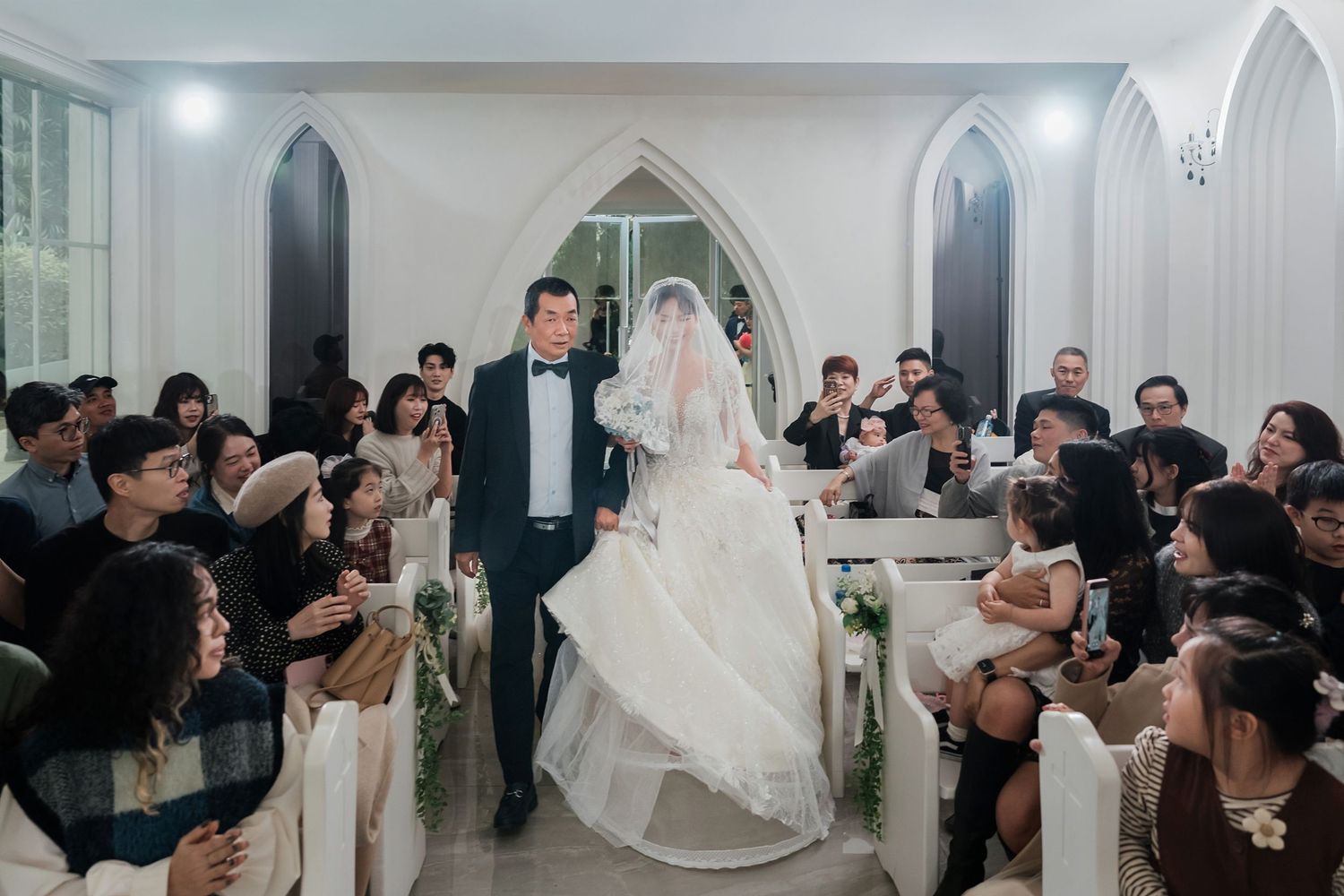 Wedding guests gather in a church during a ceremony as the bride walks down the aisle in her flowing white gown.