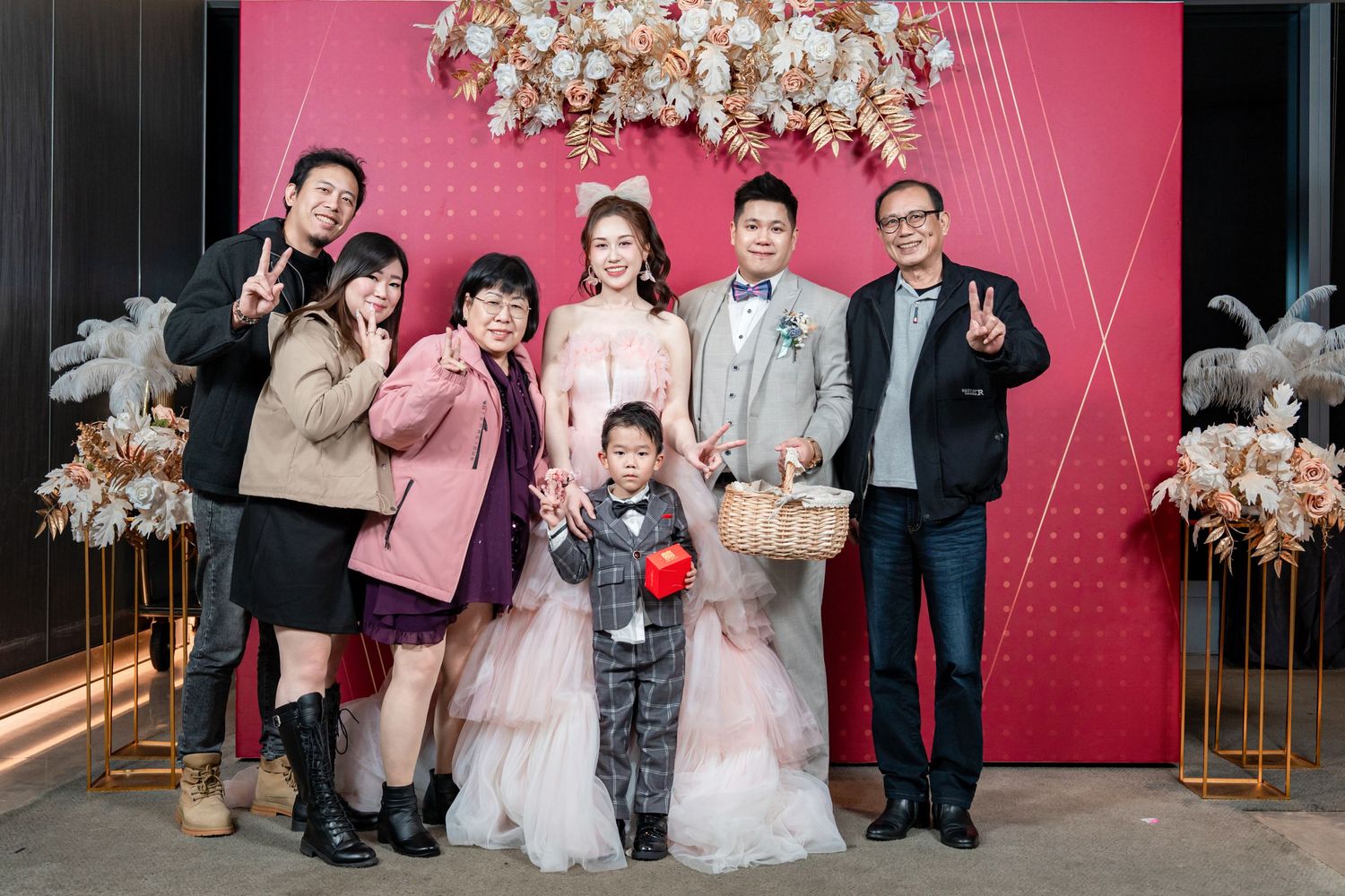 Wedding party poses together in front of pink backdrop with floral decorations at celebration event.