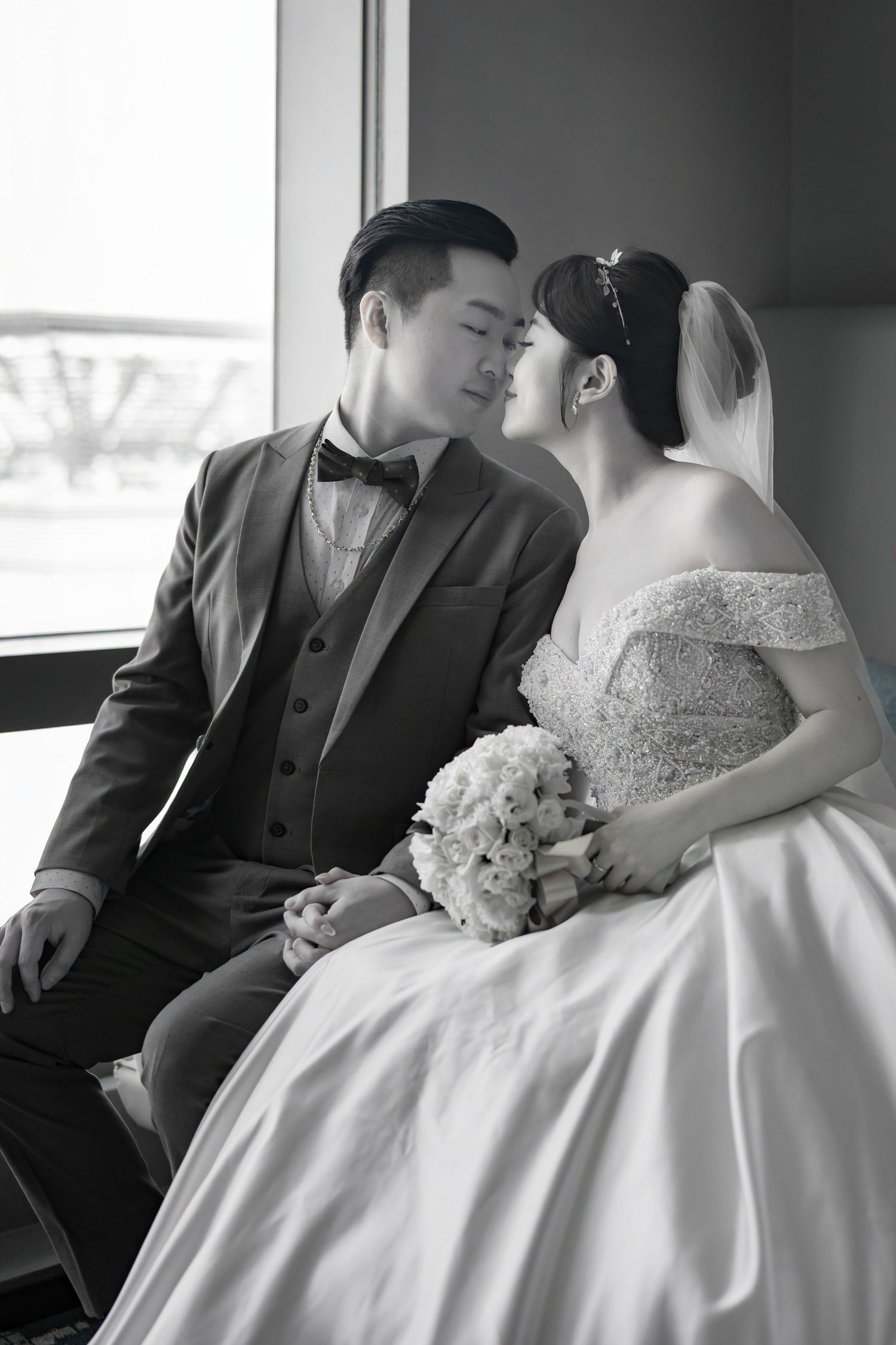 A romantic black and white wedding photo of a couple in formal attire sitting by a window with a bridal bouquet.
